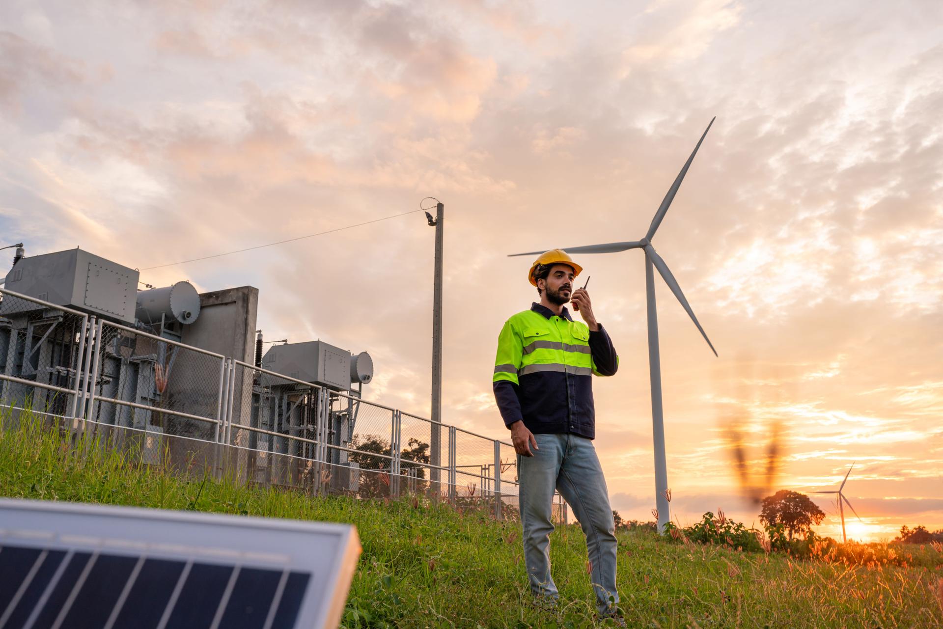 Engineer wearing uniform holding solar panels and inspection work in wind turbine farms at sunset rotation to generate electricity energy.